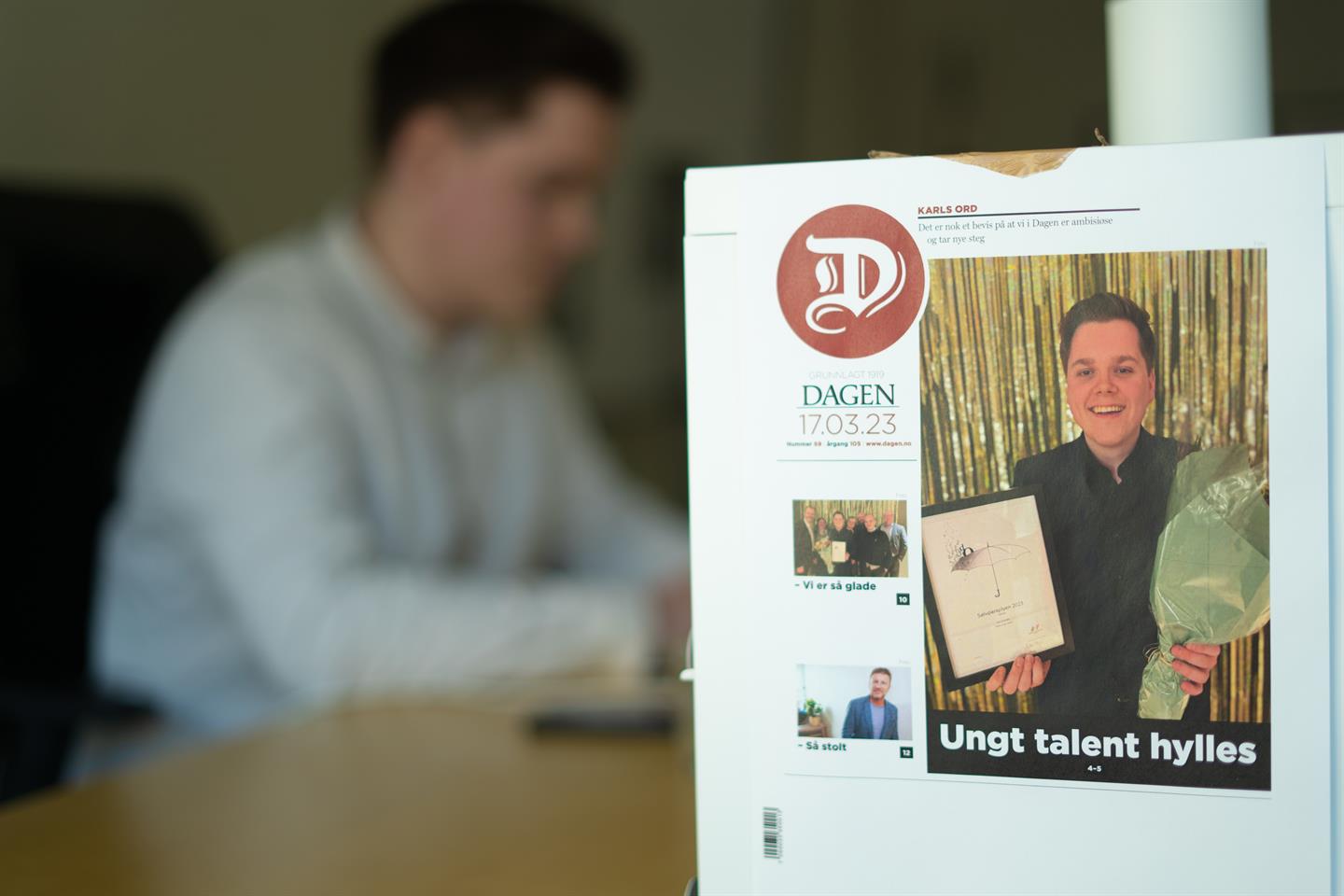 Karl Almedal jobber ved skrivepulten sin, mens det i forgrunnen henger en kopi av forsiden til Dagen med bilde av Almedal da han fant prisen for «Årets journalisttalent» i Bergen.